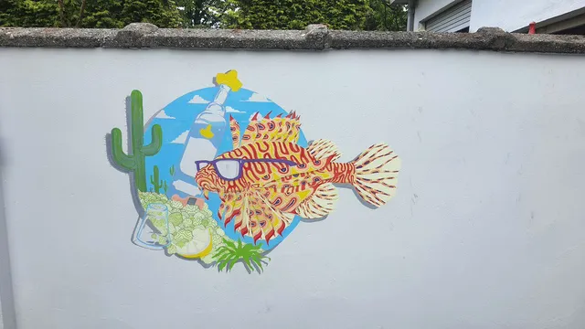Mural on a white wall featuring a stylized lionfish wearing sunglasses next to a cactus and bottle