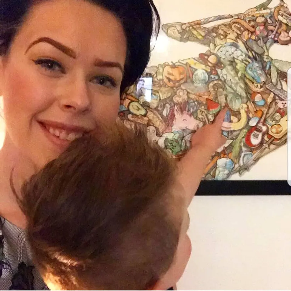 Smiling woman holding a child pointing at a framed, colorful ink illustration filled with small detailed objects