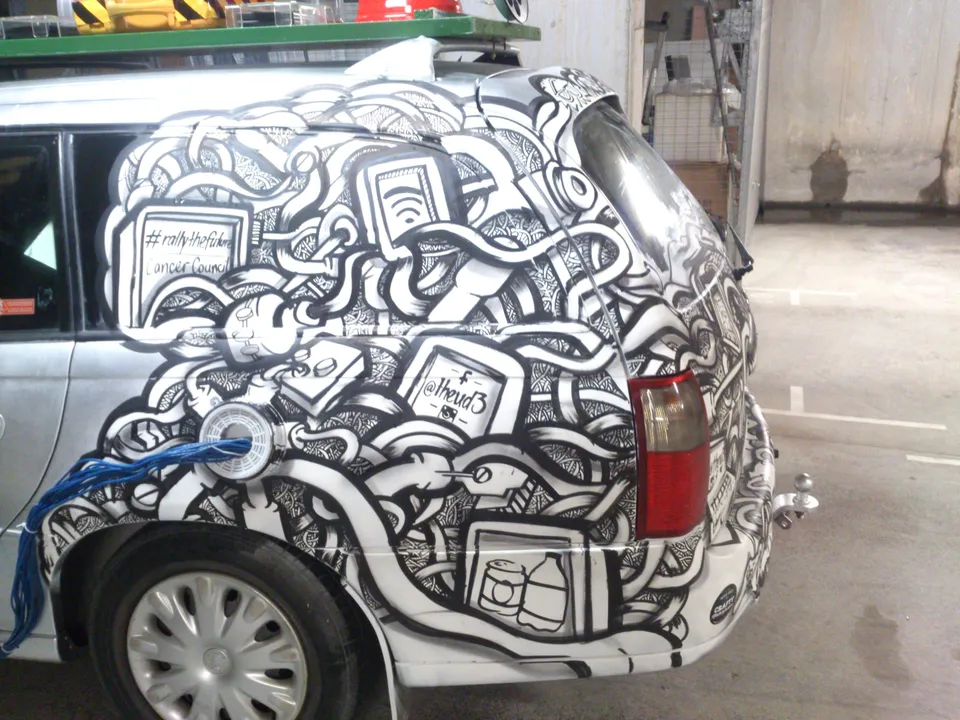 Hand-painted black and white mural of tangled cables, plugs, and screens covering the rear of a silver station wagon