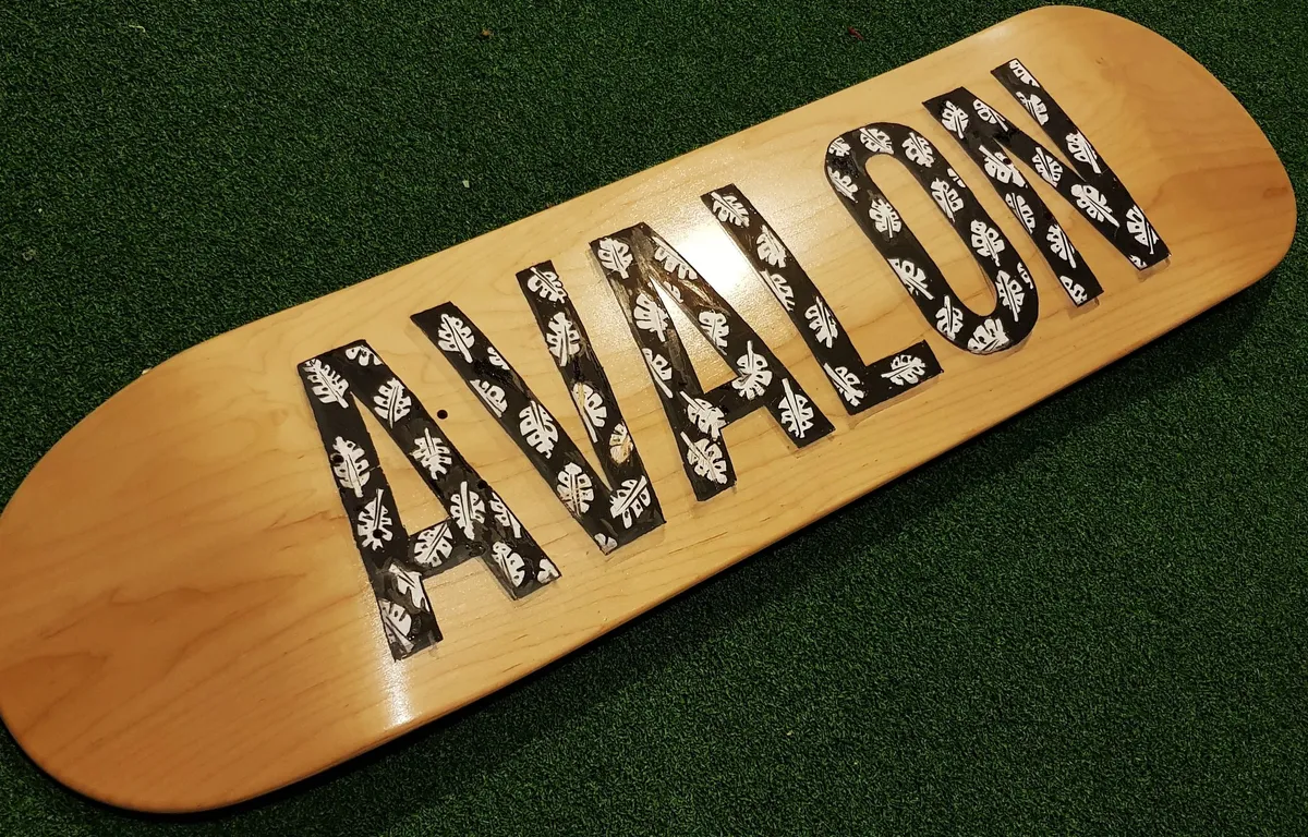 Painted wooden skateboard deck featuring the word AVALON in black letters patterned with white leaves