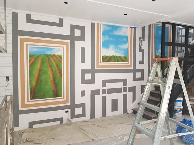 Mural in progress featuring grey geometric frames around painted scenes of green crop fields under blue skies