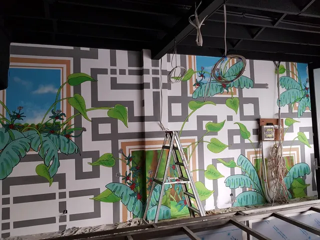 In-progress mural featuring a grey geometric maze overlaid with green tropical plants and painted sky panels