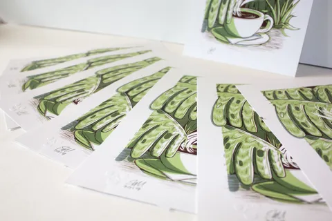 Fanned prints showing a bold illustration of a steaming coffee cup surrounded by green jungle leaves