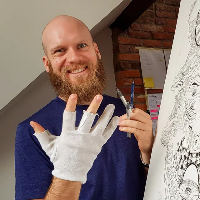 Smiling bearded artist wearing a fingerless drawing glove and holding pens beside a detailed black ink illustration