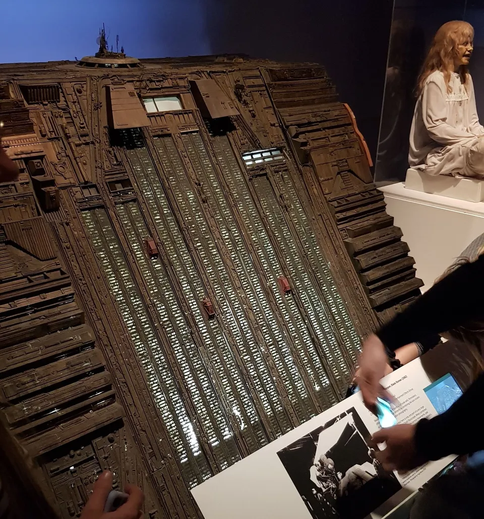 Hands illuminate an exhibition plaque next to a large sci-fi model and a pale girl mannequin