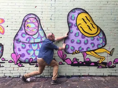 Brick wall mural of two psychedelic purple spotted figures, with a man posing as if pushing one