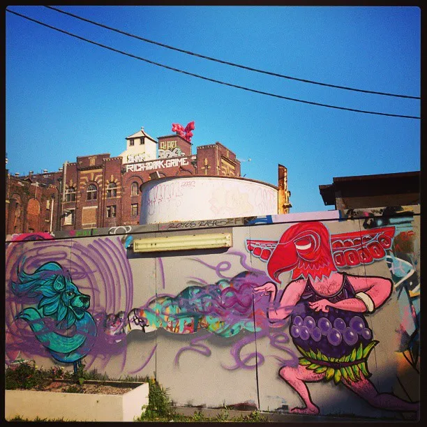 Psychedelic mural of a pink bird-masked figure with a grape torso facing a floating blue lion head in purple smoke