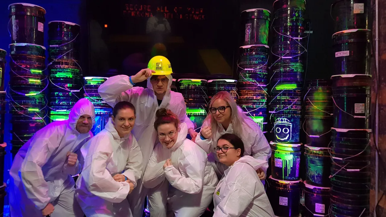 Six people in white suits pose in a blacklight room with stacks of glowing paint buckets