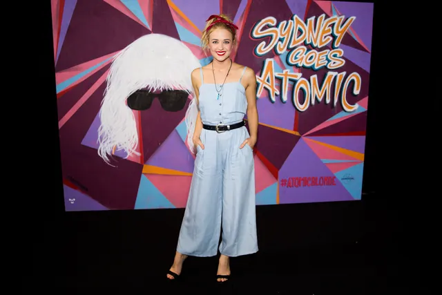 Woman posing by a geometric mural backdrop painted with a white wig, sunglasses, and the text Sydney Goes Atomic