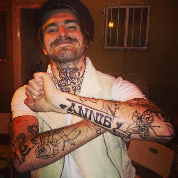 Smiling man with a mustache crossing his arms to show off bold black ink tattoos on his forearms and an owl on his neck
