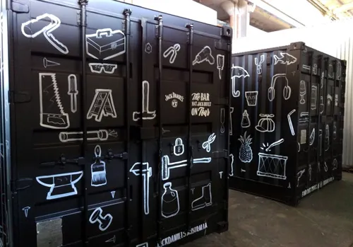 Hand-painted black shipping containers with white line art of tools, party items, and Jack Daniel's branding