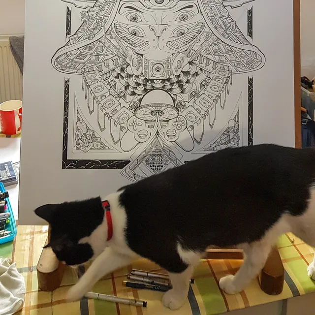 Black and white cat walking past a complex ink drawing of a symmetrical mask with speakers and game controllers