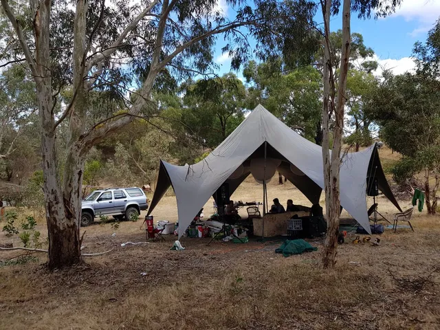 Large star-shaped canopy tent in a dry wooded area with people resting underneath near a parked silver SUV