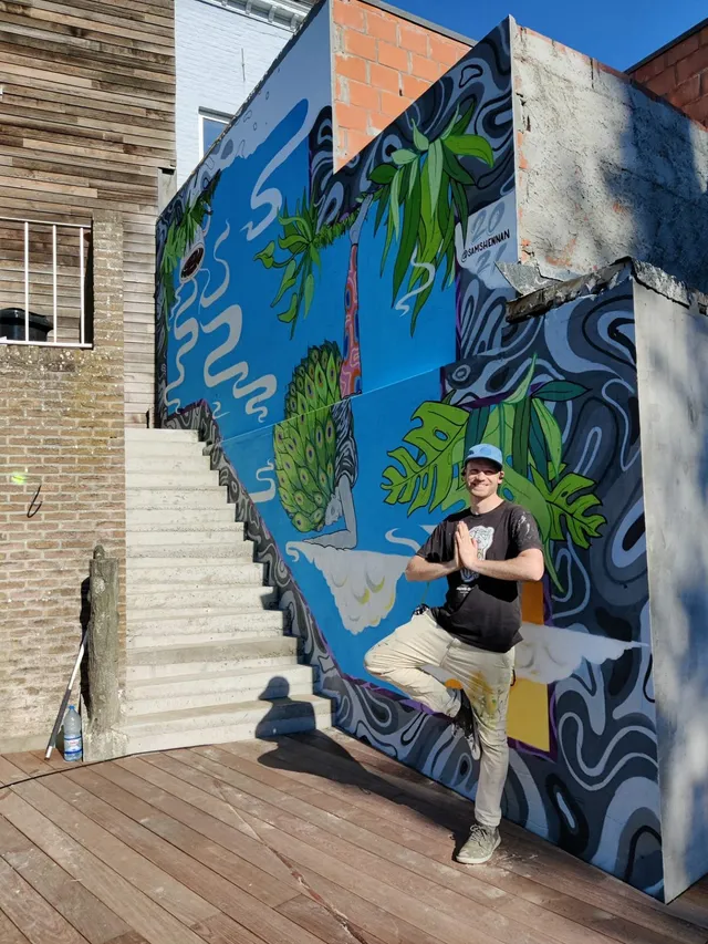 Man in a yoga pose in front of a blue outdoor mural depicting a handstanding figure with peacock feathers