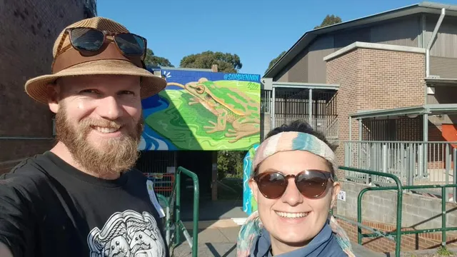 Smiling man and woman taking a selfie in front of an outdoor mural of a large green frog on a leaf