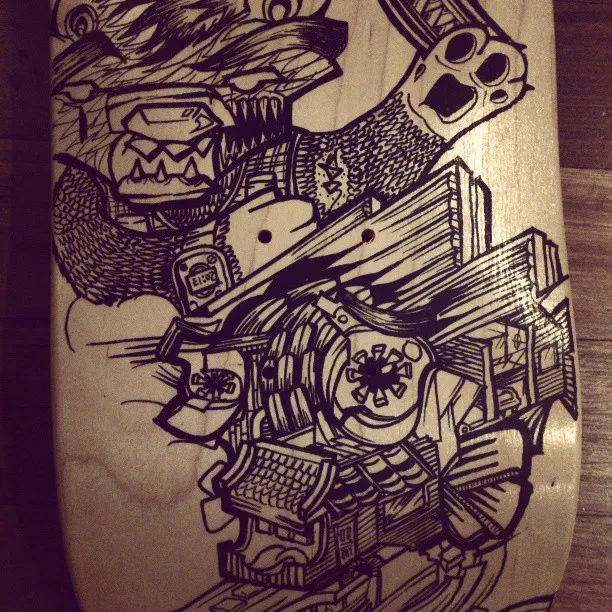 Black ink drawing on a wooden skateboard deck featuring a snarling beast, a spray can, and a traditional house