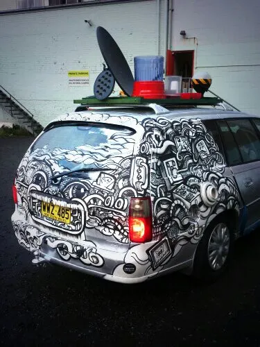 Silver station wagon hand-painted with black and white psychedelic line art featuring eyes and teeth, with a loaded roof rack