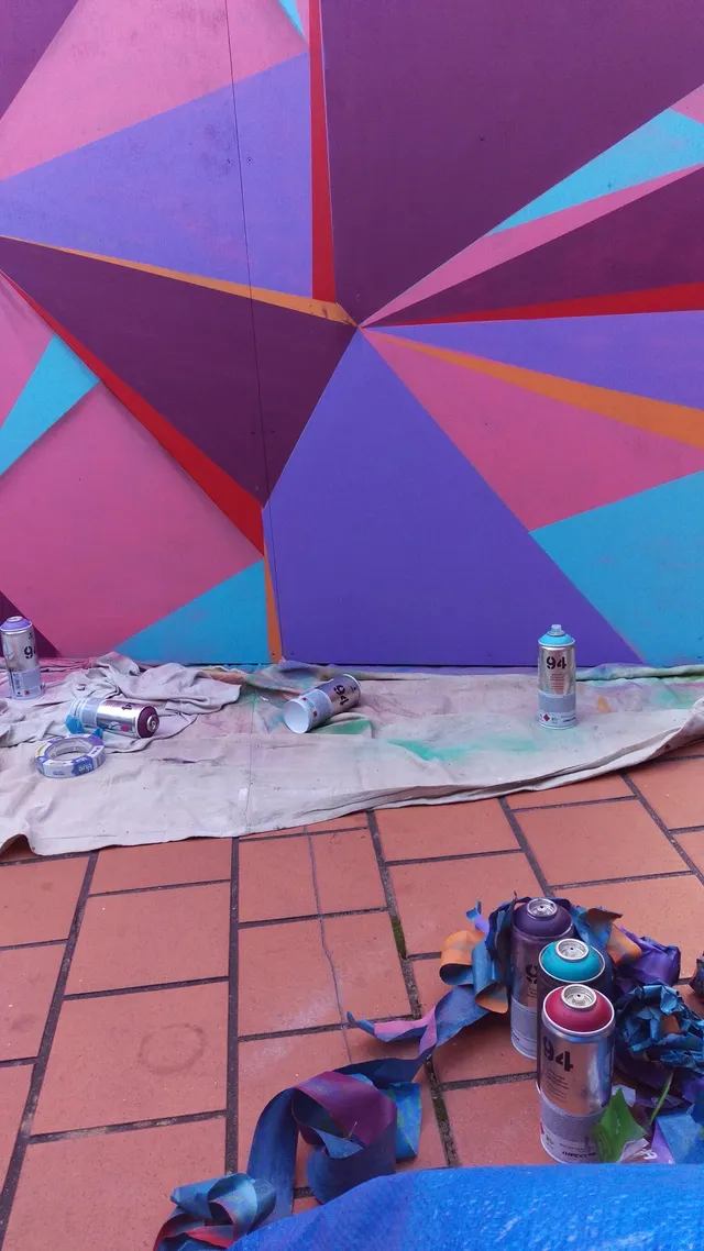 In-progress geometric mural in pink, purple and blue with spray paint cans and tape scattered on a drop cloth below