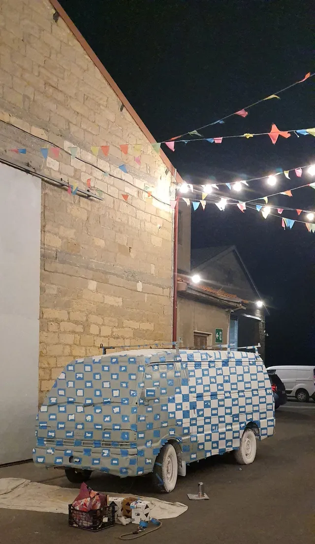 Van covered in a grid of blue painter's tape alongside painting supplies outdoors at night under string lights