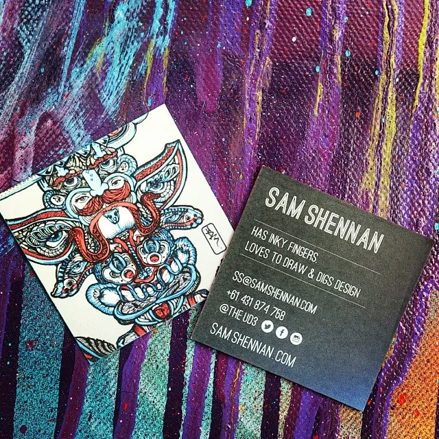 Printed square business cards on a painted background, one showing a psychedelic totemic face and the other contact info