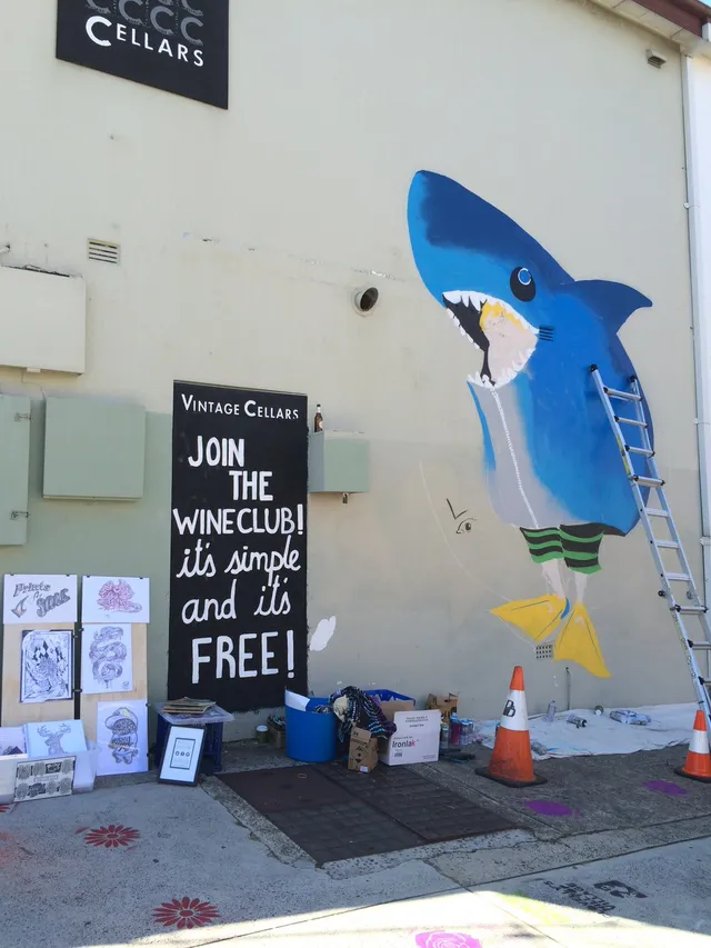 In-progress mural on an exterior wall depicting a boy in a blue shark suit and yellow flippers, next to a ladder