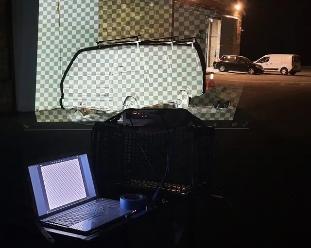 Laptop and projector setup at night casting a checkerboard van outline onto a building wall