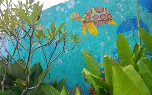 Mural on a bright teal wall showing a colorful, patterned sea turtle and bubbles, framed by real green tropical plants