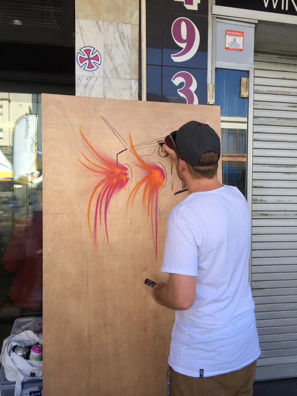 Artist drawing black linework over vibrant orange and pink spray-painted shapes on a standing wooden panel outdoors