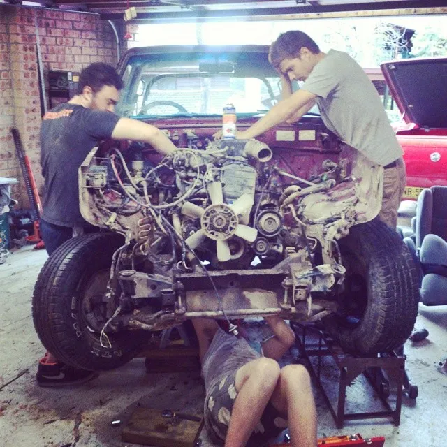 Three men working on an exposed vehicle engine in a garage, with one lying underneath the chassis