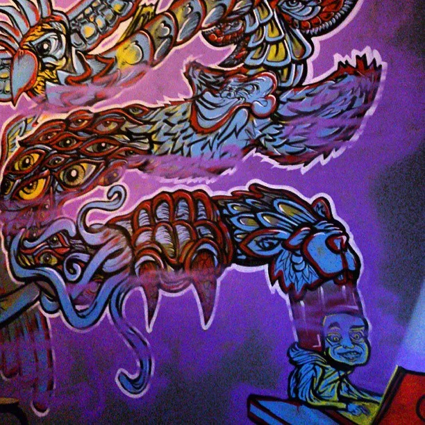 Mural of a winding psychedelic creature with animal heads and multiple eyes emerging from a blue figure on a laptop