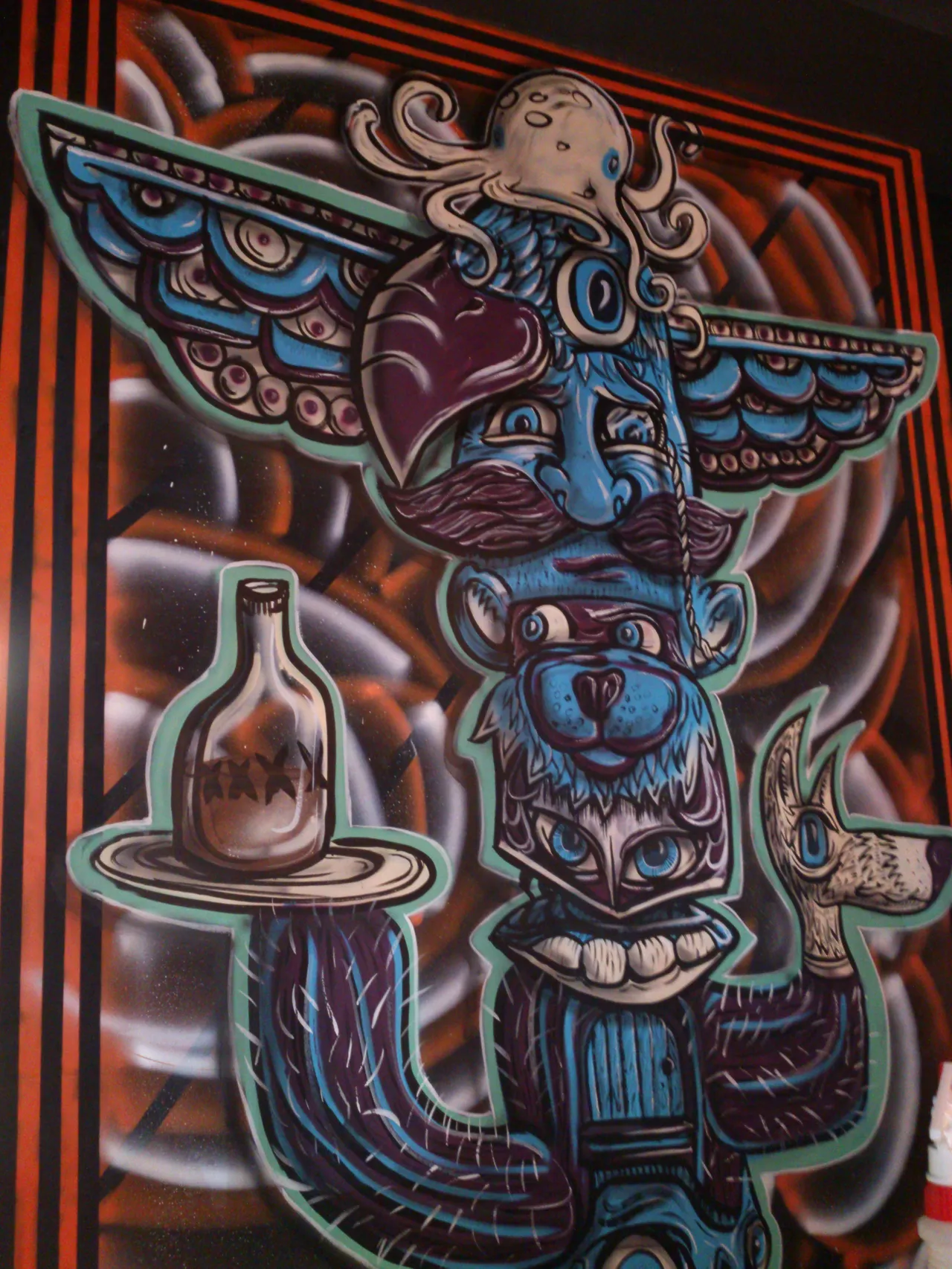 Mural of a surreal blue totem pole stacked with an octopus, mustached face, bear, and an arm holding a bottle on a tray