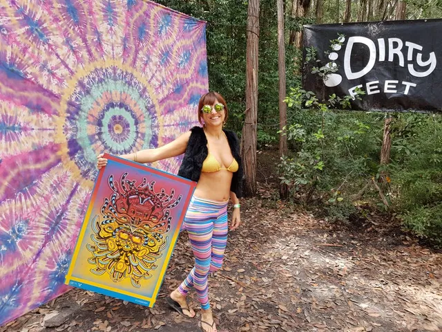 Woman holding a psychedelic poster illustration of totemic masks outdoors next to a Dirty Feet banner
