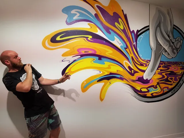Man posing playfully beside a vibrant mural of a large grey hand splashing psychedelic liquid towards him