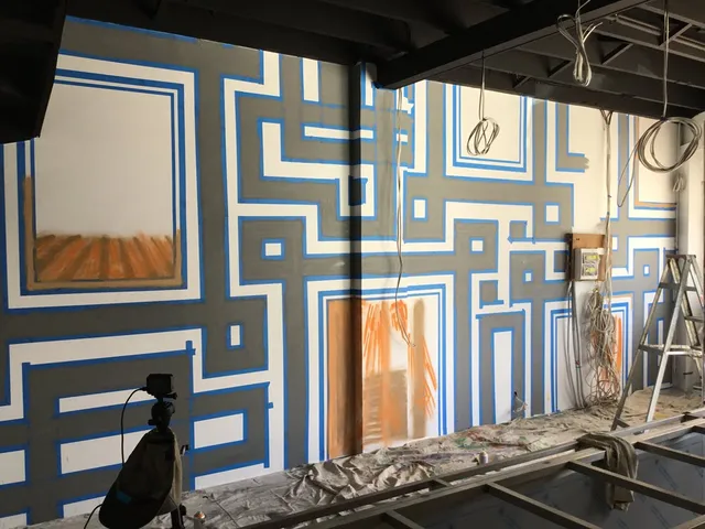 In-progress interior mural featuring a geometric pattern of thick grey lines outlined with blue painter's tape