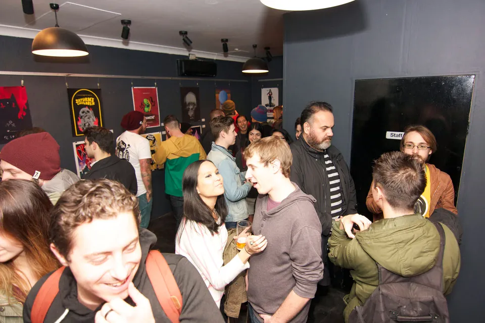 Crowd of people mingling at a gallery event with framed posters hanging on the dark walls in the background