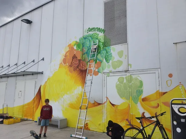 In-progress mural of a splashing yellow wave and green hops on a white building, with a ladder and two people in front