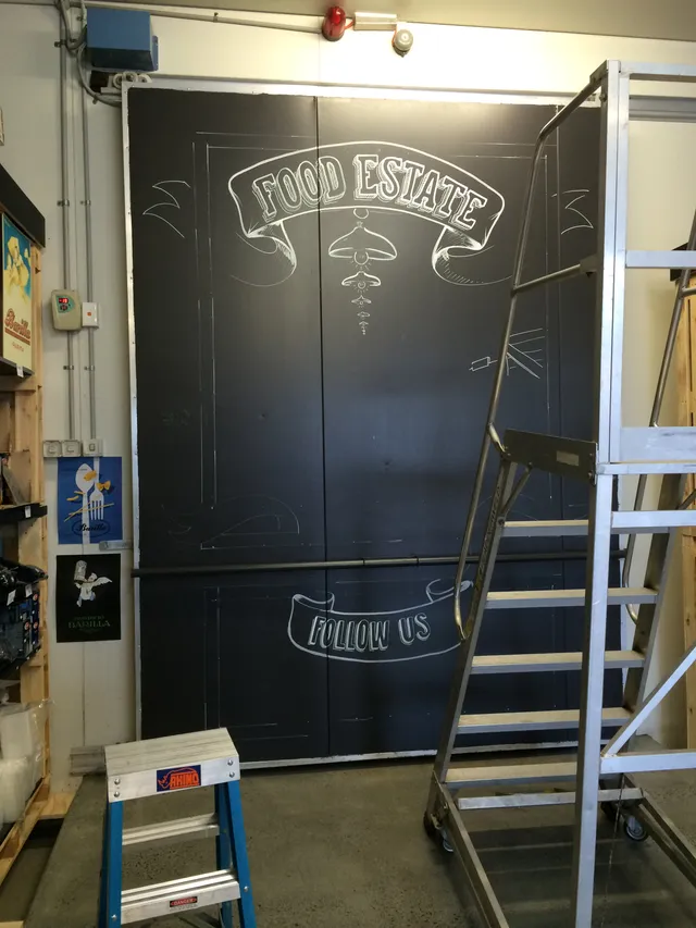 Work-in-progress mural on a black wall with white sketched banners reading FOOD ESTATE and FOLLOW US