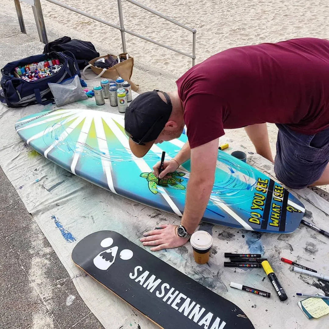Artist drawing a sea turtle on a blue surfboard with a paint marker, alongside a custom black skateboard deck