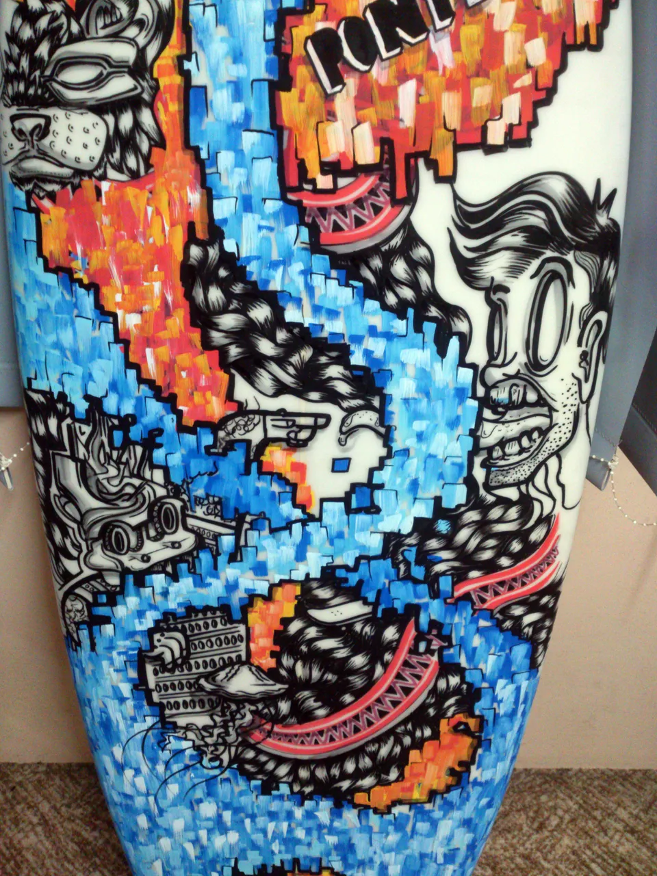 Painted surfboard with black line art of surreal faces and masks amidst pixelated blue and orange color blocks