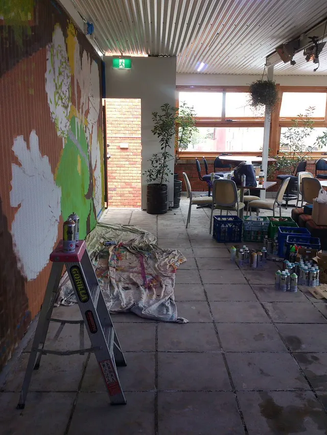 Indoor mural in progress on a corrugated wall with a stepladder, drop cloth, and spray paint cans