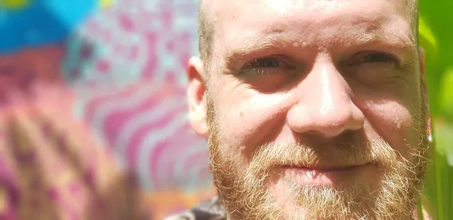 Smiling bearded man in bright sunlight standing in front of a blurred, colorful abstract mural