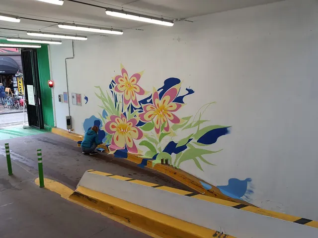 Person painting a vibrant mural of large pink and yellow flowers on the white wall of a carpark ramp