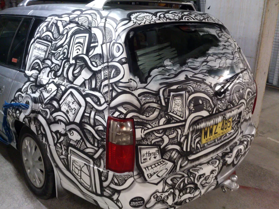 Silver car covered in a black and white hand-painted mural of bold intertwining lines, cables, and tech objects