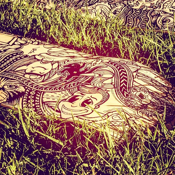Hand-painted skateboard deck in grass featuring intricate black line art of a smiling face and swirling totemic patterns