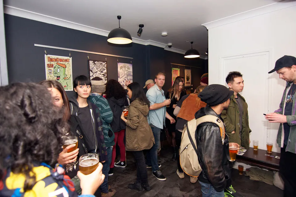 Crowd of people mingling and holding drinks at an exhibition with illustrated posters hanging on a dark wall