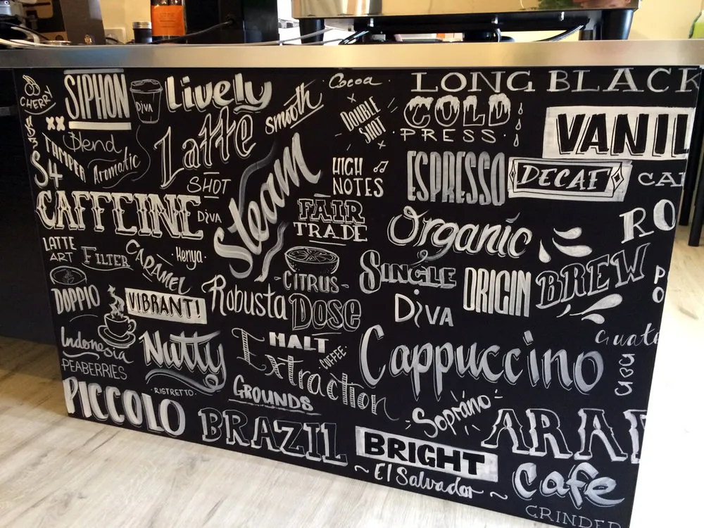 Hand-painted mural on a black cafe counter featuring diverse white typography of coffee terms