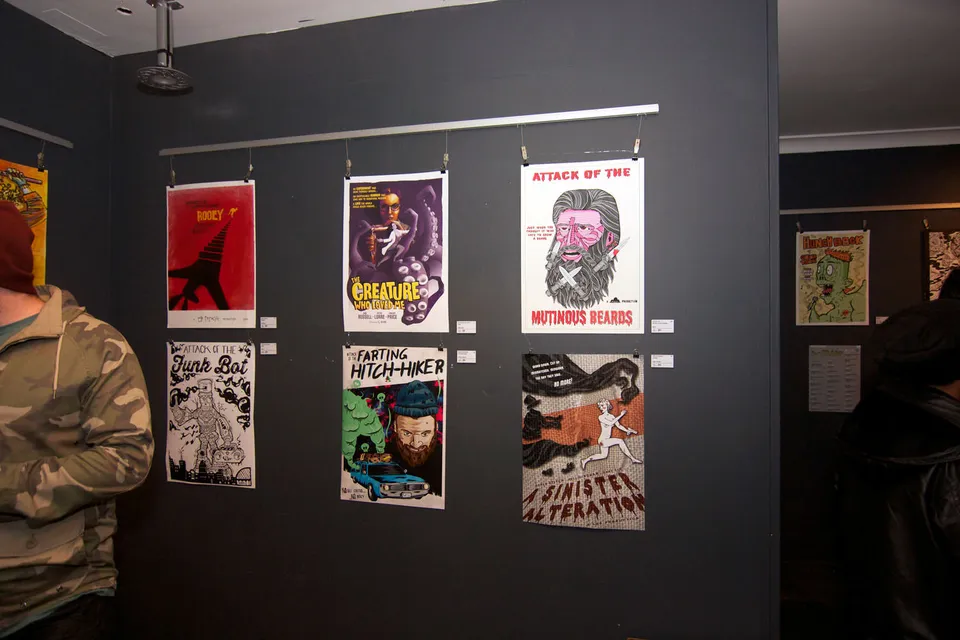 Six illustrated B-movie posters hanging from clips on a dark gallery wall at an exhibition