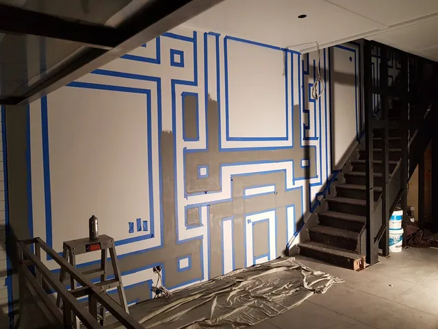 Work-in-progress mural showing blue painter's tape outlining thick geometric grey lines on a white interior wall