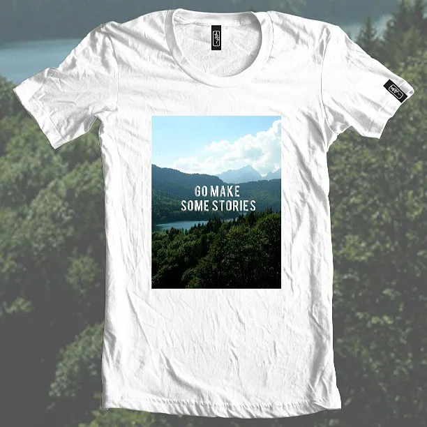 White t-shirt printed with a forest and mountain landscape and the text GO MAKE SOME STORIES