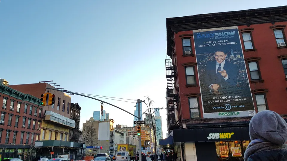 City street with a large billboard for The Daily Show with Trevor Noah on a red brick building